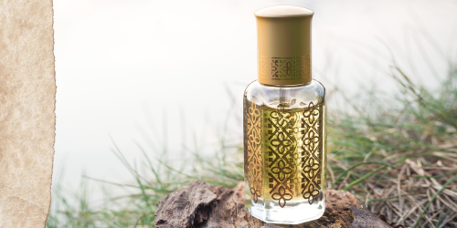 Hellgelber Attar in mit Gold verzierter Glasflasche vor natürlichem hellen Hintergrund und Gras.
